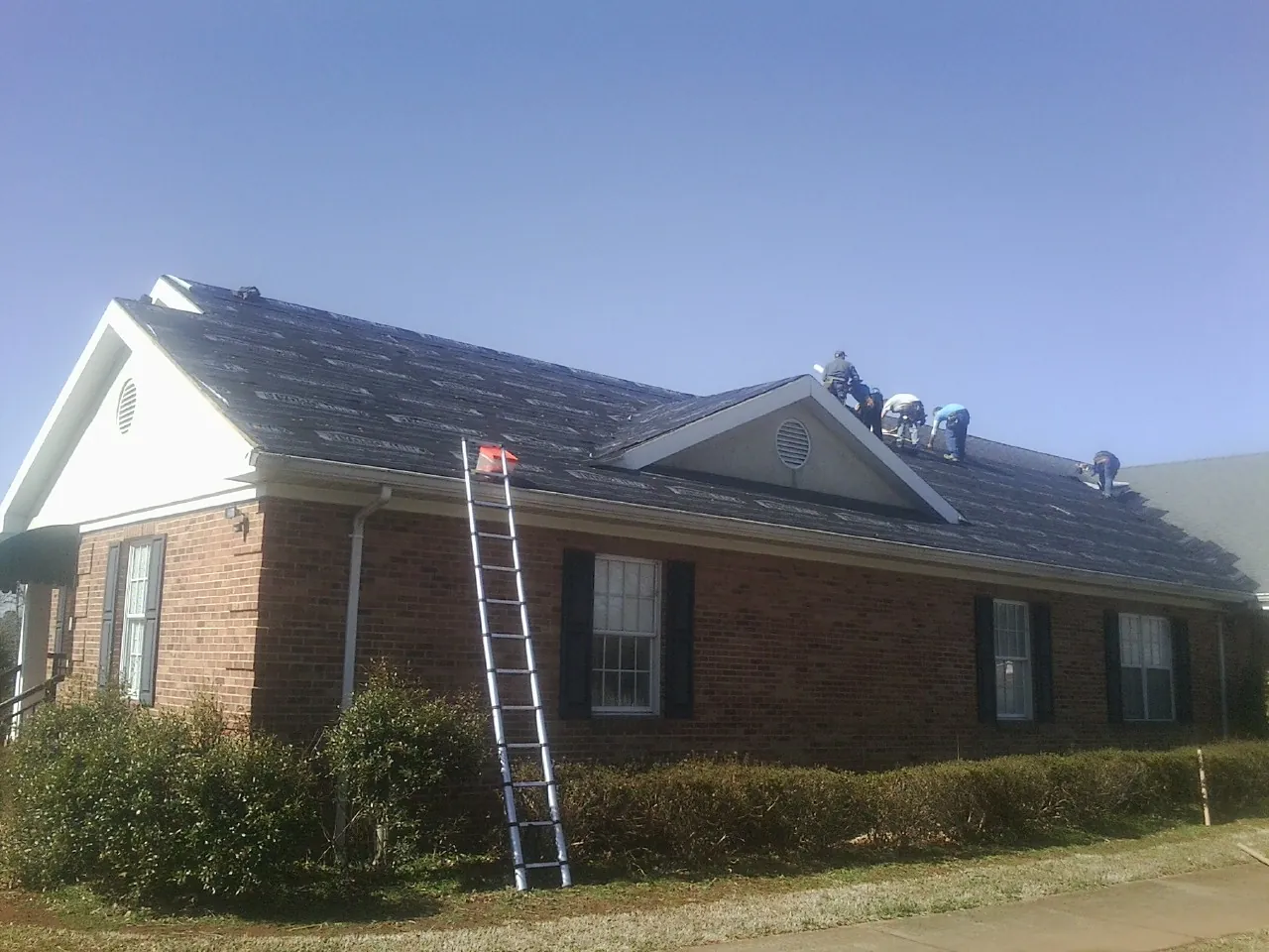 Crew tearing shingles off a brick ranch roof with a ladder set — Metal Roof Repair work by our crew in Roberts